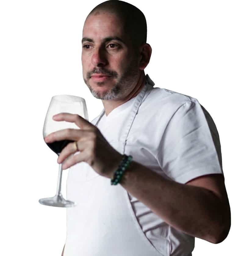 השף אבירם דותן לבוש בבגדי עבודה בהירים, מחזיק כוס יין בידו ומביט הצידה. צילום פורטרט נקי על רקע בהיר המשדר מקצועיות, רוגע וחיבור לאירוח ולקולינריה.