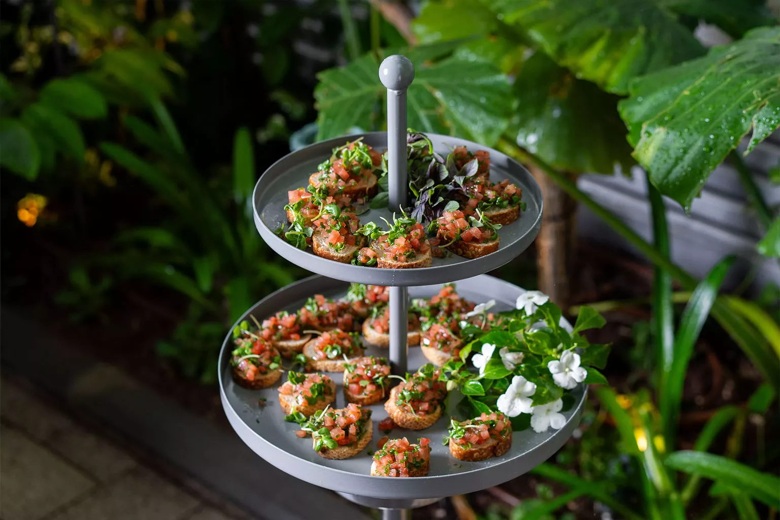 מגש הגשה דו-קומתי עם קרוסטיני קטנים ועליהם תערובת עגבניות קצוצות, עשבי תיבול וירק טרי. המגש מוצב בסביבה ירוקה עם צמחייה, ומדגיש אירוח קליל ורענן.